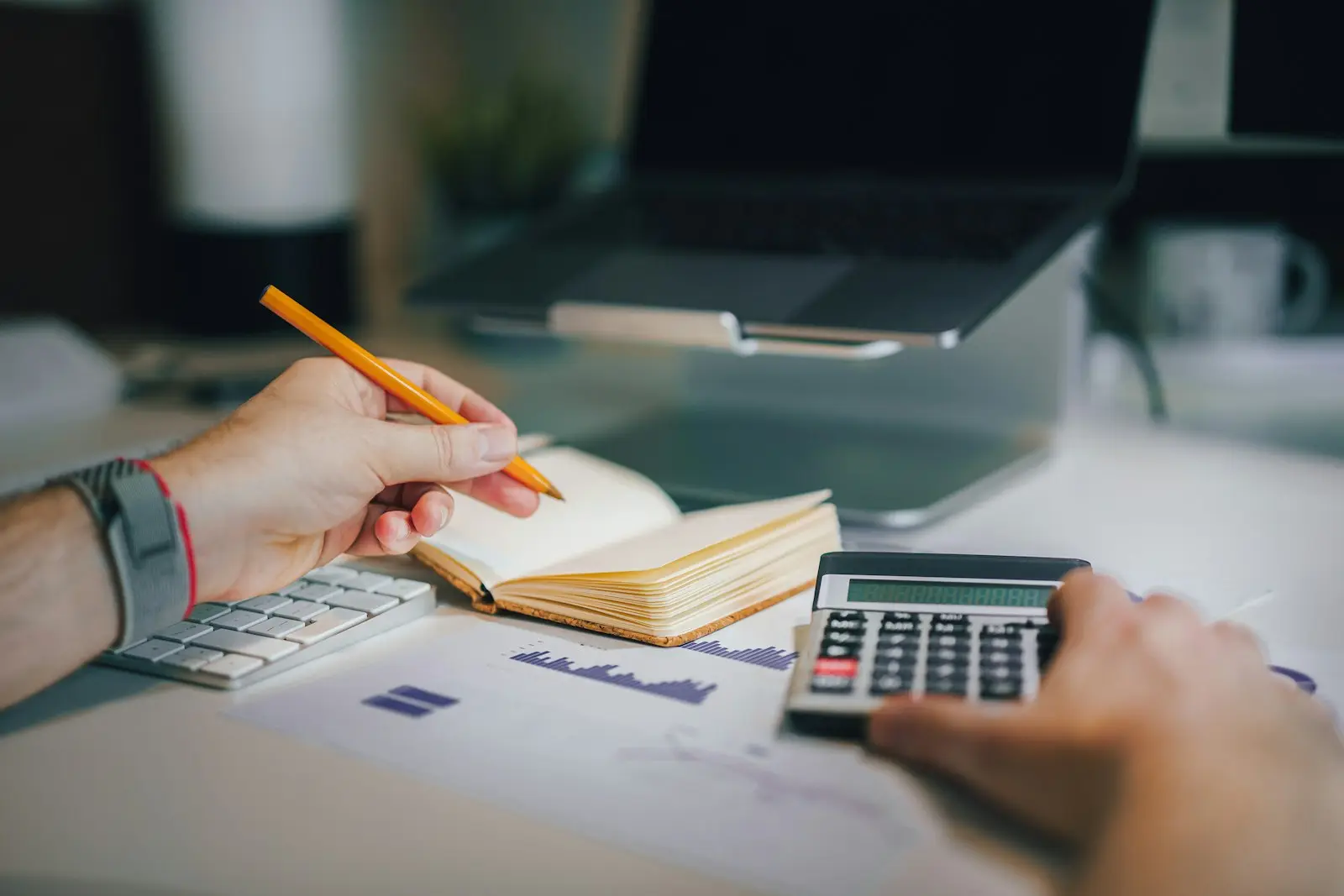 Mains tenant un crayon et une calculatrice devant un carnet et un ordinateur portable